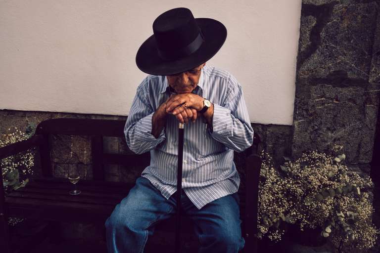 Photo de rue en couleur d'un vieil homme bien habillé, avec un chapeau noir et une canne. Il est assis sur un banc en pleine reflection à Cordoue, en Espagne.