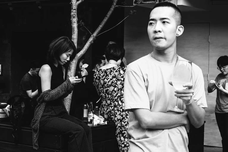 Photo de rue en noir et blanc d'un homme tenant un verre de vin blanc, avec une femme en arrière plan qui regarde son téléphone portable à Taipei, Taiwan.