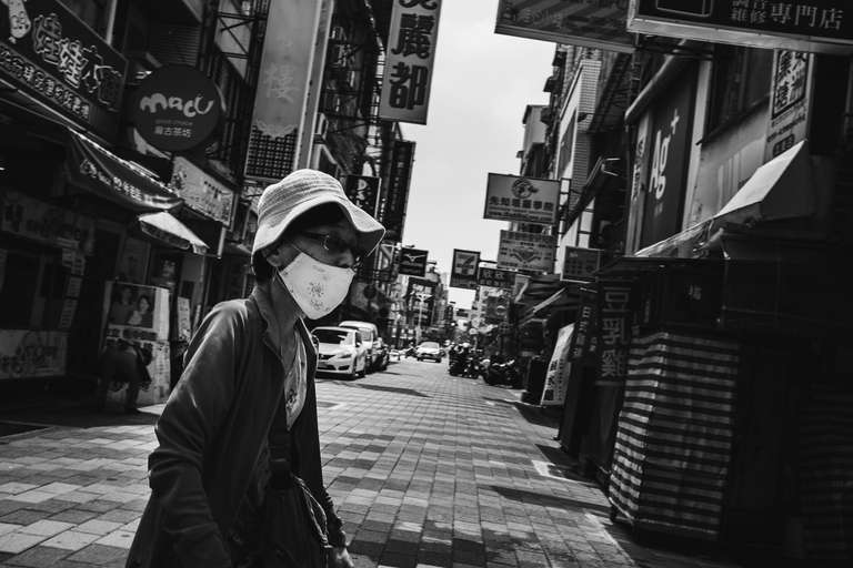 Photo de rue en noir et blanc d'une femme âgée portant un masque et un chapeau dans une rue ensoleillé à Tainan, Taiwan.