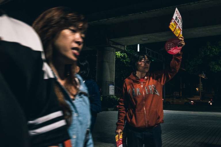 Photo de rue en couleur de nuit, un couple passe devant une femme tenant un flyer publicitaire au dessus de sa tête à Taipei, Taiwan.
