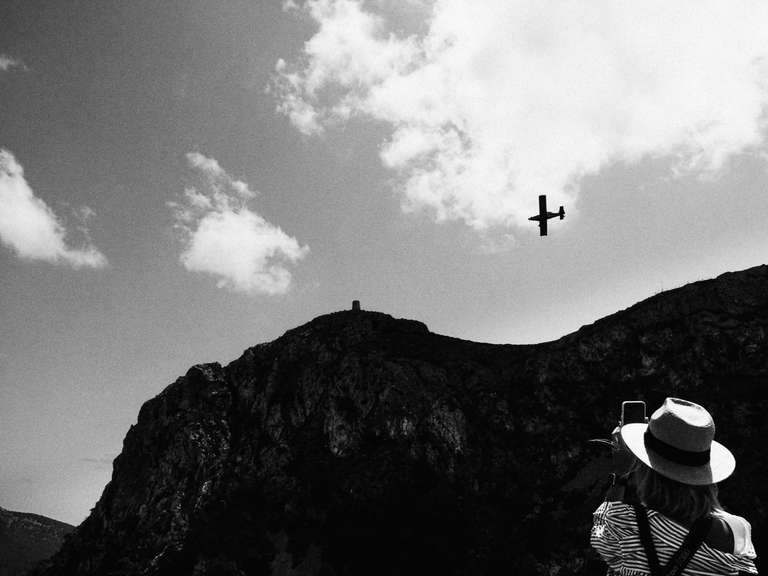 Photo de rue en noir et blanc d'une femme portant un chapeau, et prenant en photo un avion volant au dessus du cap de Formentor à Majorque, en Espagne.