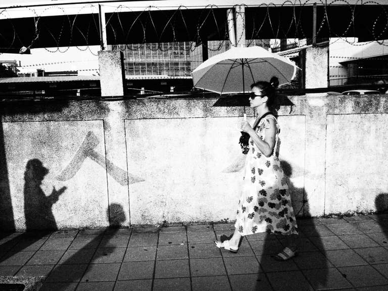 Photo de rue en noir et blanc d'une femme portant une ombrelle marchant devant un mur d'une enceinte militaire à Taipei, Taiwan.