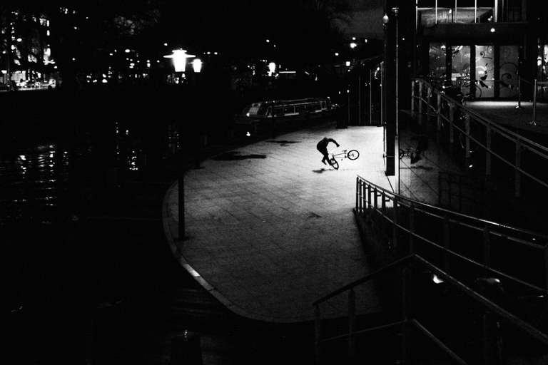Photo de rue en noir et blanc d'un homme faisant une figure de bmx, la nuit sur les quais des canaux de Amsterdam, Pays Bas.