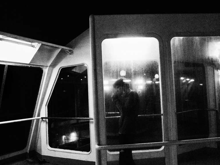 Photo de rue en noir et blanc d'un homme allumant une cigarette abrité derrière un mur vitré sur le pont supérieur d'un ferry.