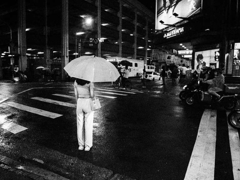 Photo de rue en noir et blanc d'une jeune femme toute vêtue de blanc, tenant un parapluie blanc, sous la pluie, au passage piéton, la nui à Taipei, Taiwan.
