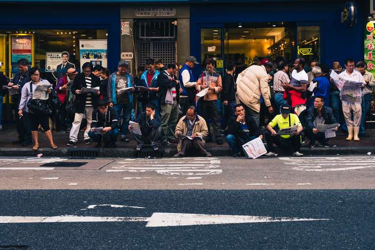 Color street photography. A picture of a crowd waiting for a bus, sitting on the floor or standing up and reading the news paper. Hong Kong.