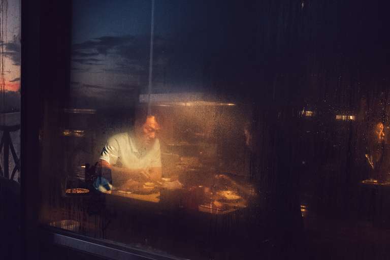 Color street photography. A picture of a couple having hot pot for dinner. They are seen through a steamy window during the sunset in Taipei, Taiwan.