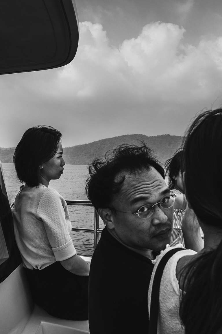 Black and white street photography. A picture of a group of people on a boat on Sun Moon Lake. One man is starring into the camera whilst a woman is looking to the horizon. Taiwan.