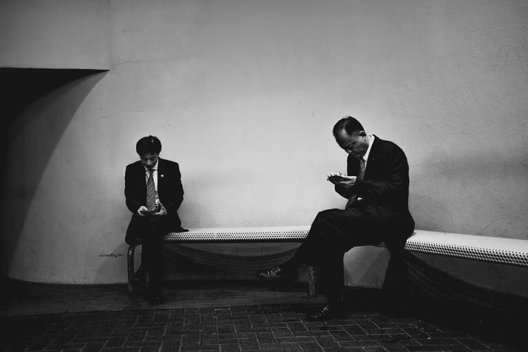 Black and white street photography. A picture of a couple of business man wearing suits on their cellphone, sitting on a bench in Hong Kong.