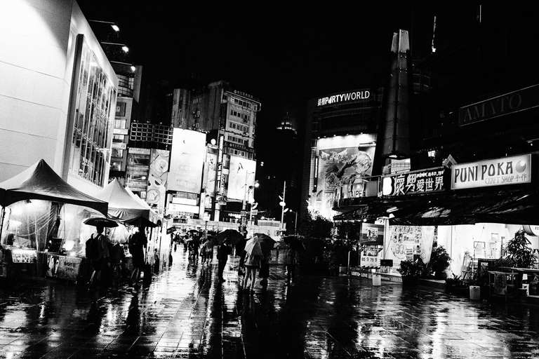Black and white street photography. A picture of a market at night, in the rain, the light from the building is reflected on the ground. Taipei, Taiwan.