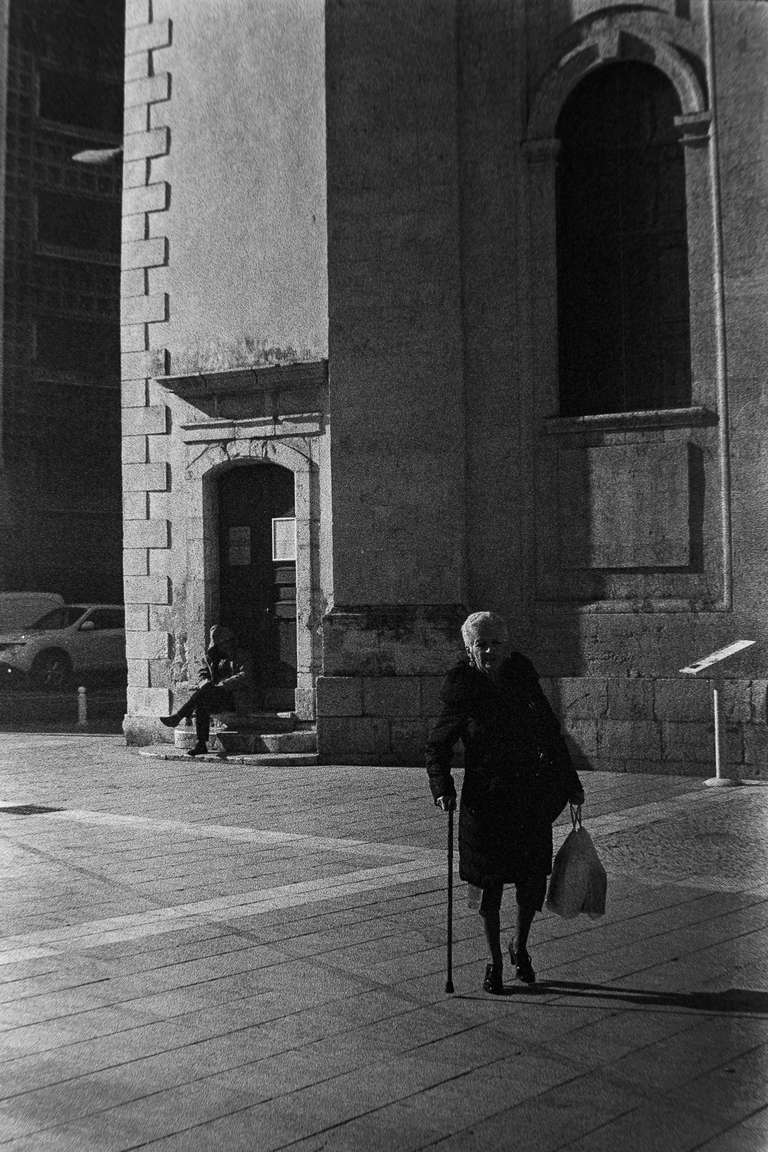 Black and white street photography. A picture of an old woman walking in front of a church, she's dress in black and she is using a cane. Toulon, south of France.