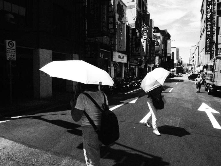 Black and white street photography. A picture of people crossing a road, they are using white umbrellas in the sun. Taipei, Taiwan.