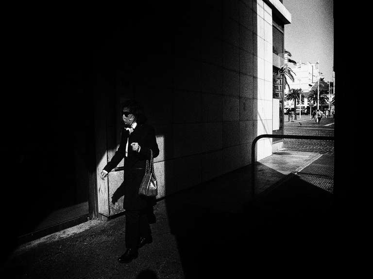 Black and white street photography. A picture of a woman walking into the sunlight, she is dressed in black and is wearing a mask. Toulon, south of France.