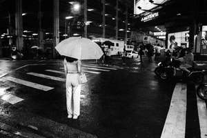 Black and white street photography. A picture of a women dressed in white and carrying a white umbrella, she is waiting at a cross road under a spotlight during the night in Taipei, Taiwan.